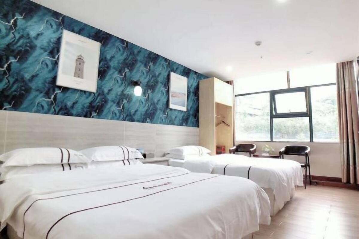 A hotel room with two beds, a desk, and a chair. There is a window with curtains, a wall with a blue abstract design, and a picture of a lighthouse.