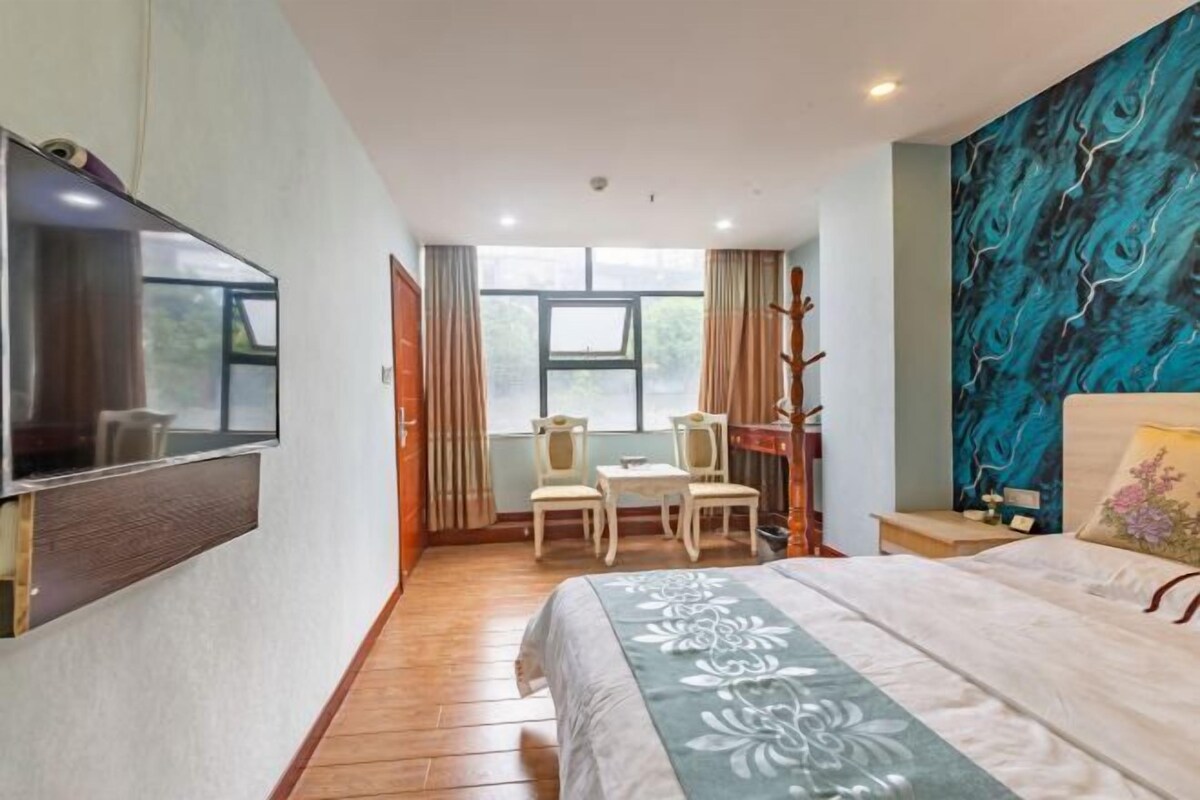 A hotel room with a bed, a desk with chairs, a TV mounted on the wall, and a large window.