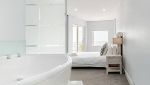 Une salle de bain moderne avec une grande baignoire, un lit avec du linge de maison blanc et une petite table de chevet avec une lampe.