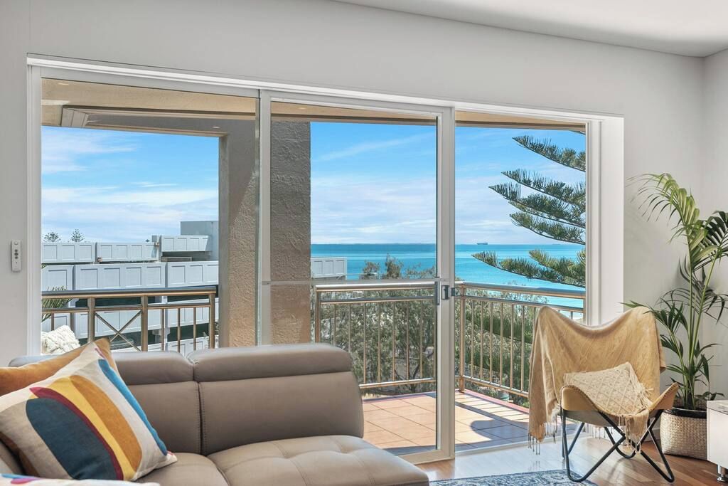 A modern living room with a sofa, a chair, and a view of the sea through large sliding doors.