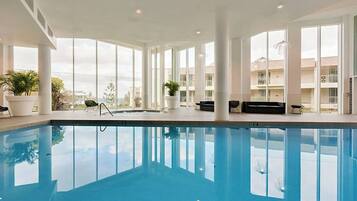 An indoor swimming pool with clear glass walls, potted plants, and a view of a building in the background.