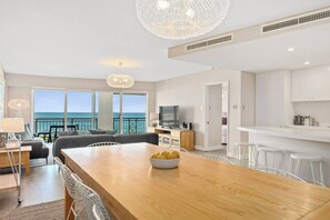 A modern living room with a wooden dining table, a sofa set, and a view of the sea through large windows. - Apartment M503 - panoramic ocean views for miles! (Scarborough)