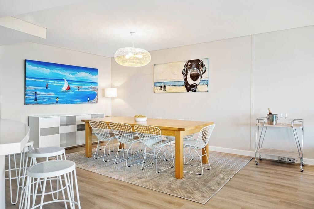 A dining area with a wooden table, white wire chairs, and a rug. There are paintings on the walls and a pendant light above the table.