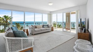 Ein geräumiges Wohnzimmer mit großem Fenster mit Meerblick, einem bequemen Sofa, einem weißen Korbstuhl und einem Flachbildfernseher.