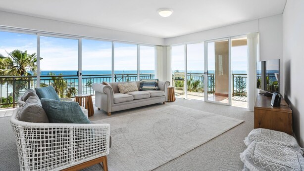 Un salón amplio con un gran ventanal con vistas al mar, un sofá cómodo, una silla de mimbre blanco y un televisor de pantalla plana.