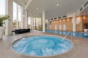 A hot tub in a spacious indoor area with large windows, a seating area, and a wooden wall with built-in cabinets.