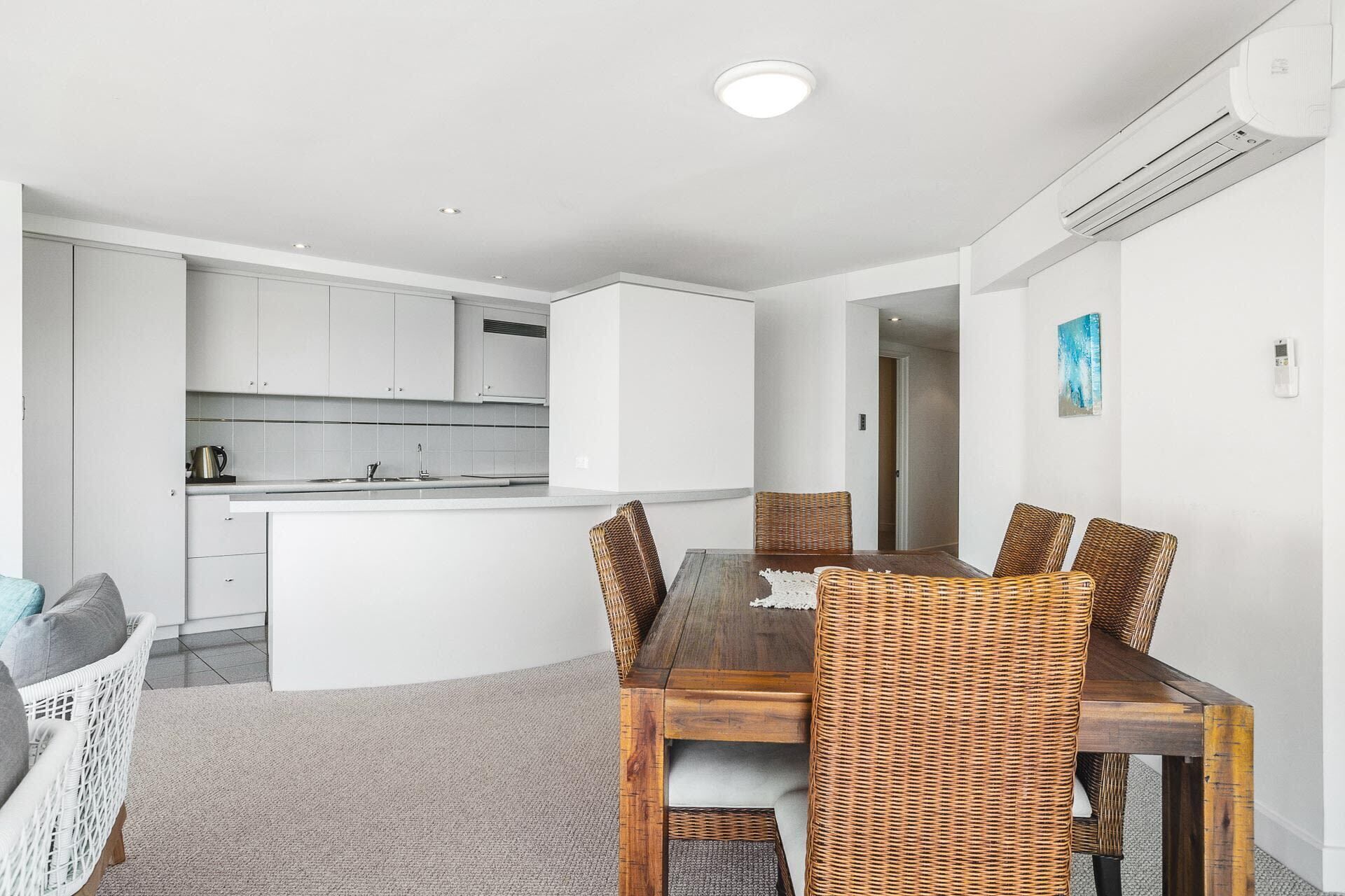 A modern kitchen with white cabinets, a dining table with wicker chairs, and a visible air conditioning unit.