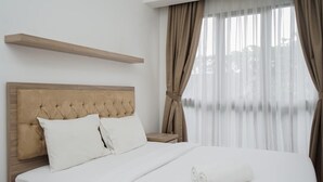 Ein ordentlich bezogenes Bett mit weißen Leinen und einem aufgerollten Handtuch, ein Holz-Kopfteil und ein Fenster mit durchscheinenden Vorhängen.