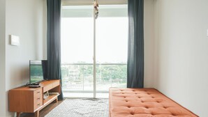 A bedroom with a large window, a wooden dresser, a television, and a tufted orange bed. - Nice And Homey 1Br At Citralake Suites Apartment (Jakarta)