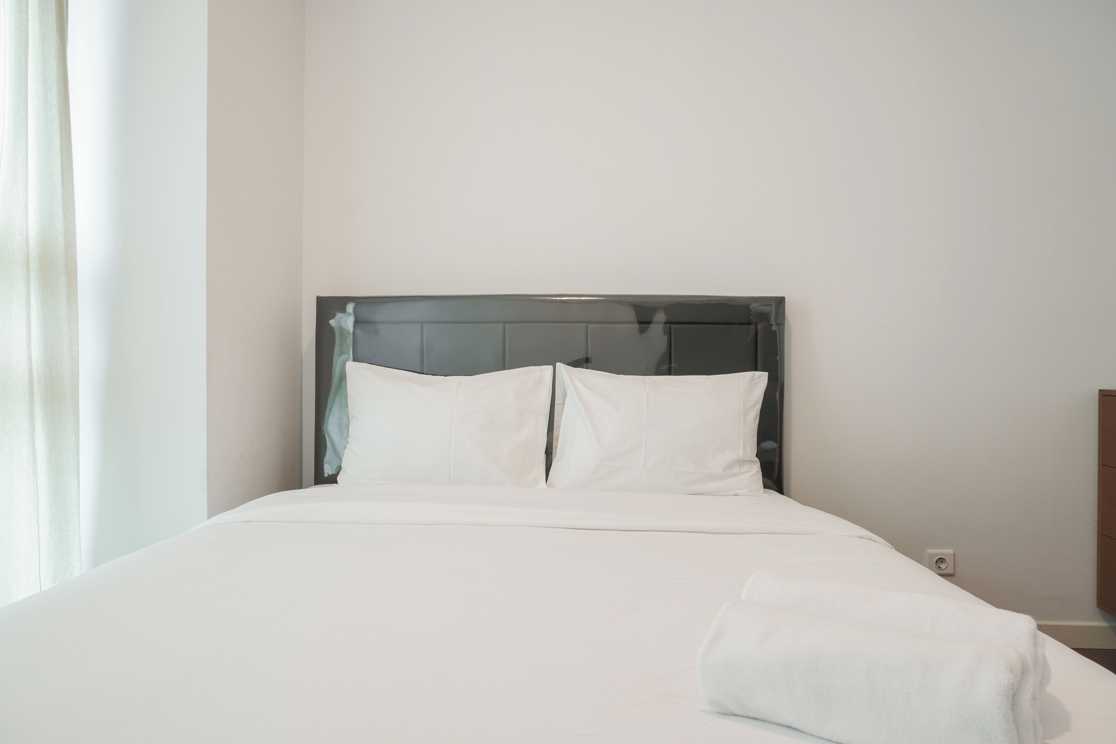 A neatly made bed with white linens and pillows, a wooden headboard, and a white wall.