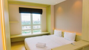 A hotel room with a bed, a bedside table, a window with a view of the city, and a built-in bench under the window. - Comfortable And Modern Look 1Br At Citralake Suites Apartment (Jakarta)