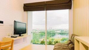 A modern living room with a television on a white stand, a brown sofa, a wooden chair, and a large window offering a city view. - Comfortable And Modern Look 1Br At Citralake Suites Apartment (Jakarta)
