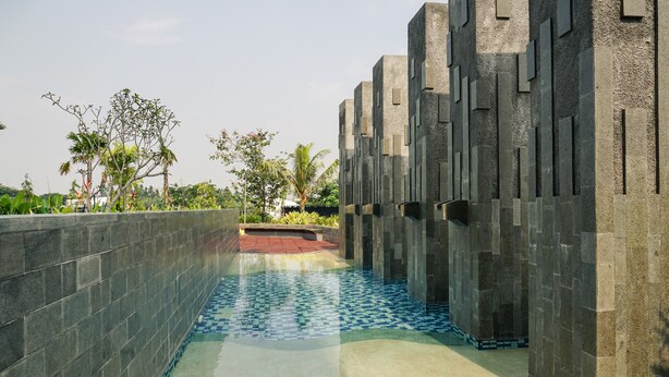 A pool area with tiled flooring, a low wall, and a pathway leading to a building with a geometric facade. - Comfortable And Modern Look 1Br At Citralake Suites Apartment (Jakarta)