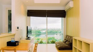 A modern living room with a sofa, a dining table with chairs, a TV, and a view of a cityscape through large windows. - Comfortable And Modern Look 1Br At Citralake Suites Apartment (Jakarta)