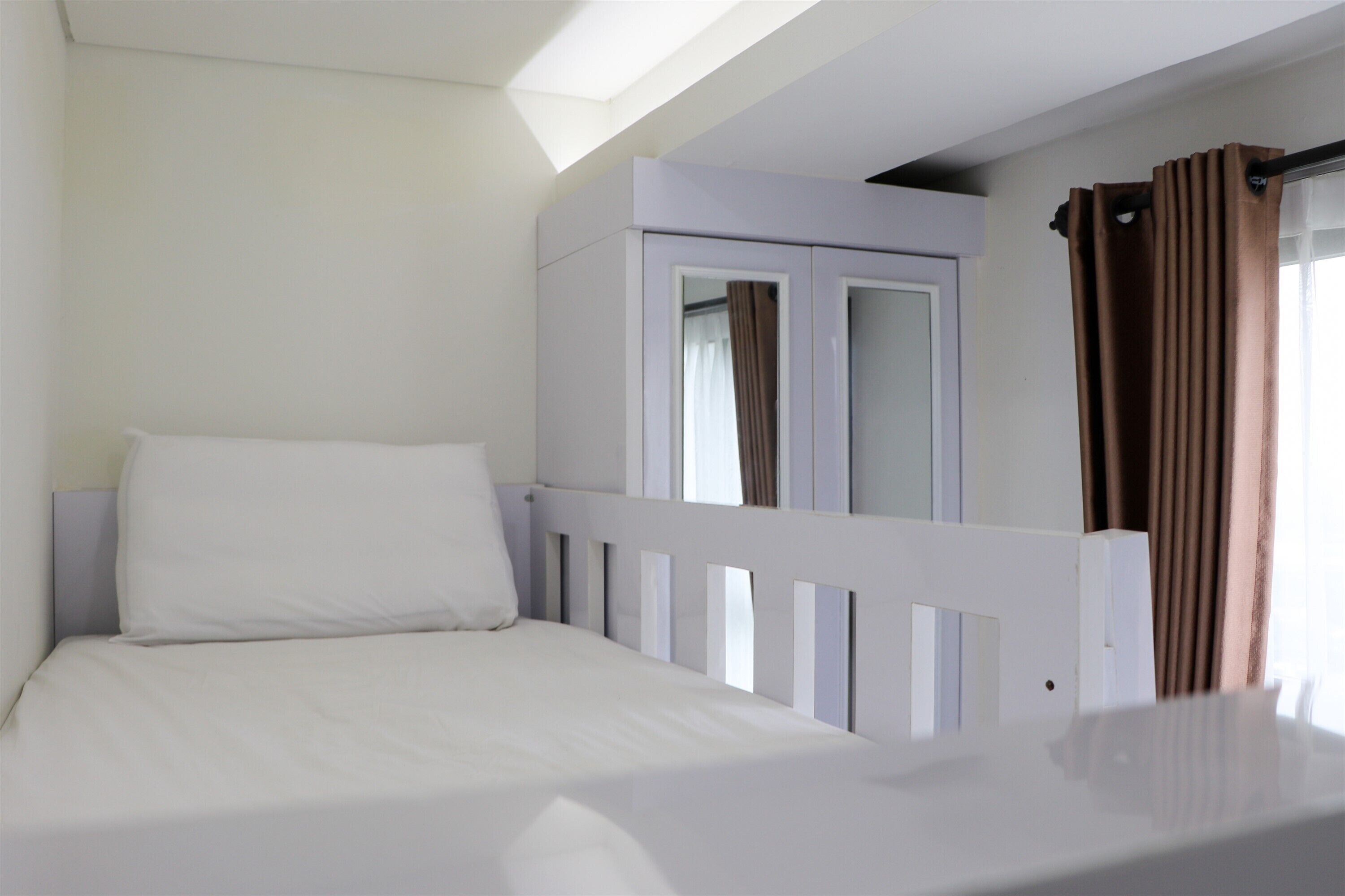 A single bed with a white headboard and a white mattress in a room with a sloped ceiling and a window with brown curtains.