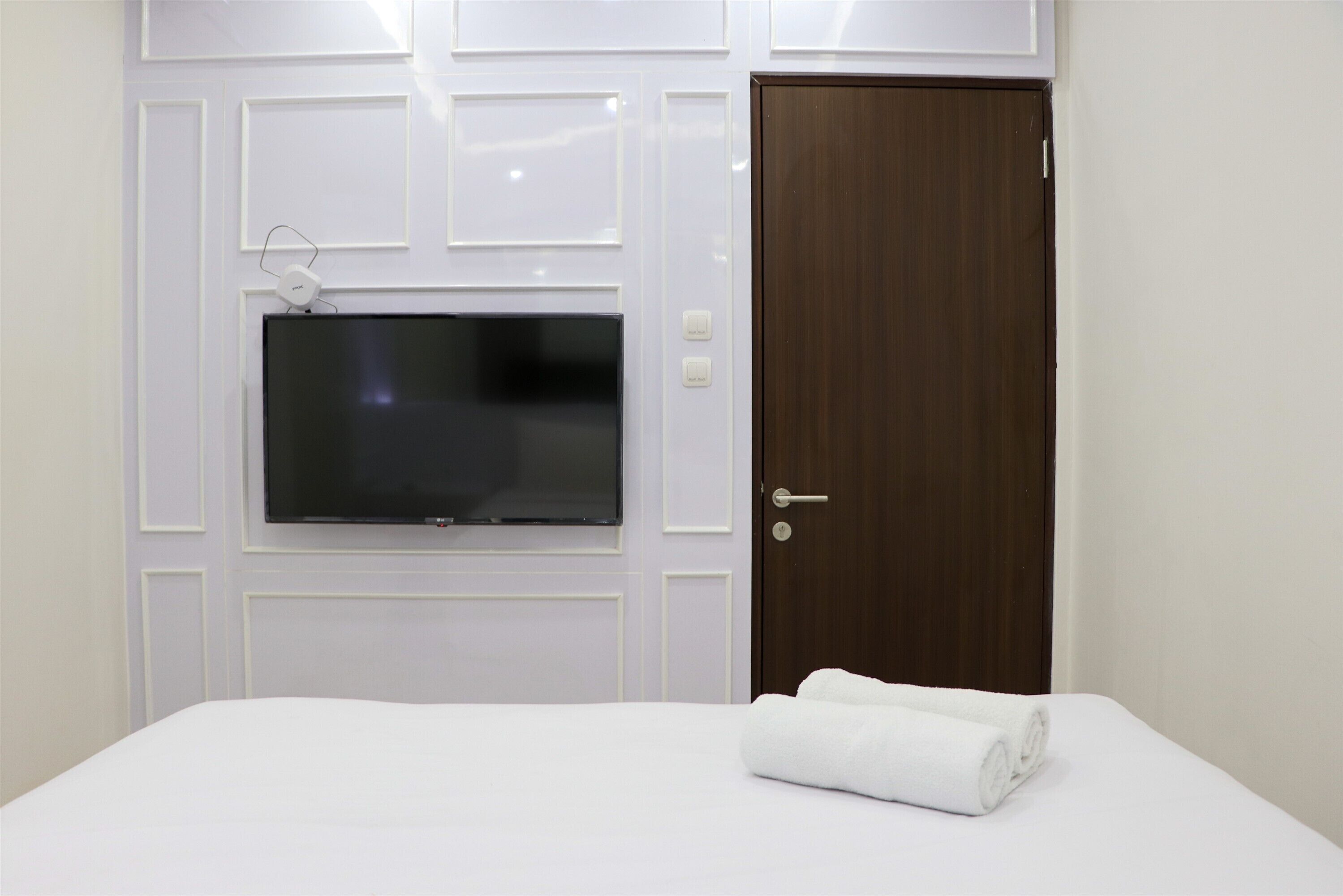 A hotel room with a flat-screen TV mounted on the wall, a wooden door, and a neatly made bed with a rolled towel.