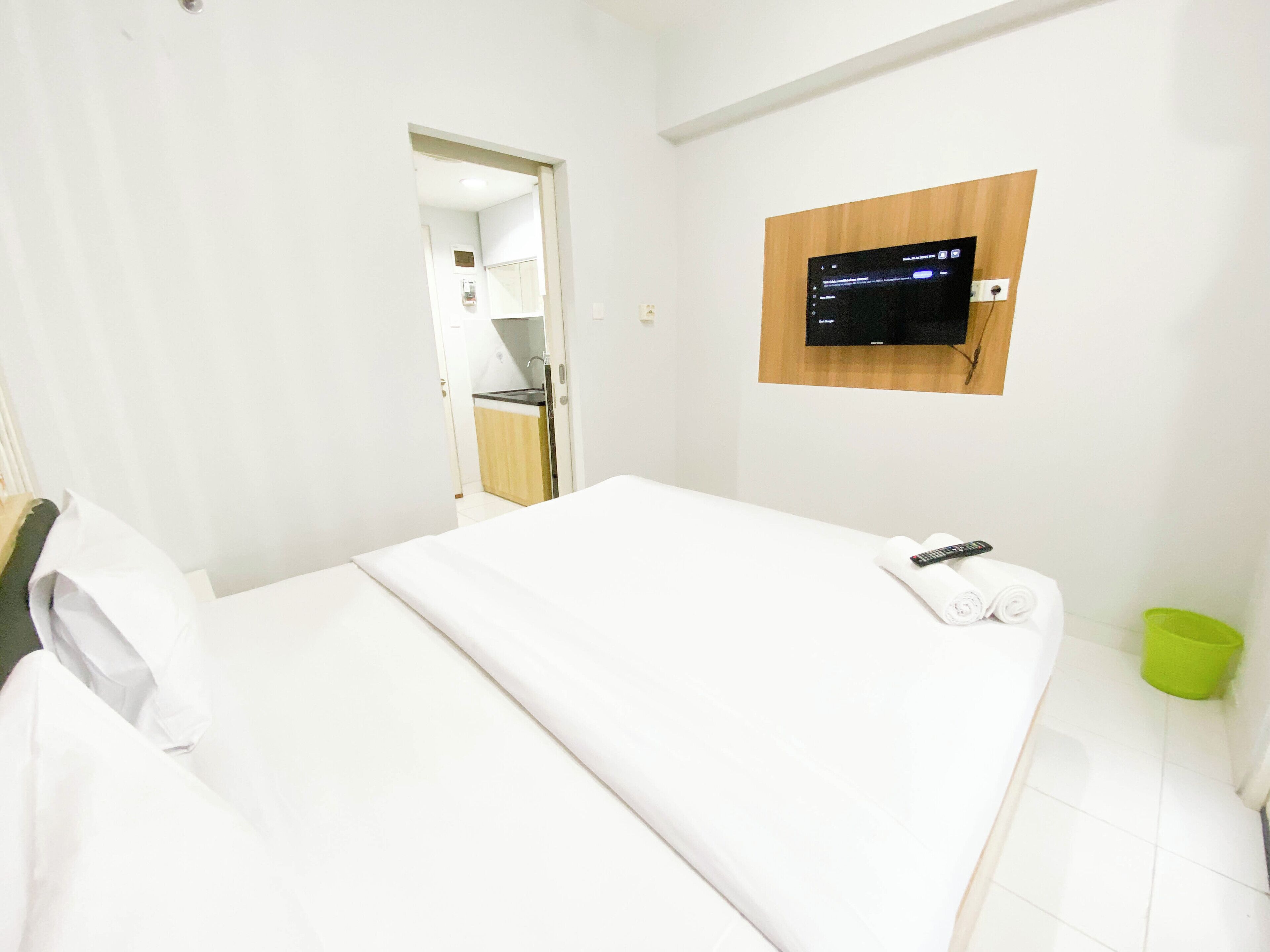 A hotel room with a bed, a TV mounted on the wall, a green trash bin, and a door leading to a bathroom.