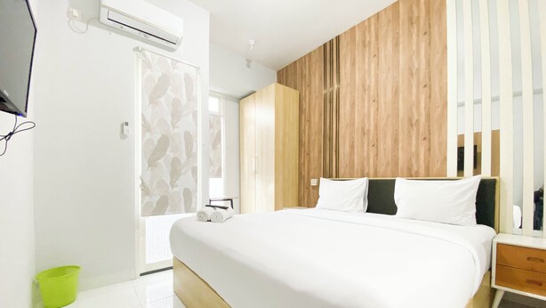 A modern hotel room with a large bed, a wooden accent wall, and a bathroom with a shower curtain. - Homey 1Br Without Living Room At Amartha View Apartment (Semarang)