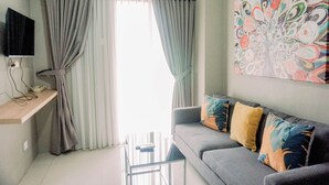 A modern living room with a grey sofa, colorful cushions, a flat-screen TV on a wall-mounted shelf, and a large window with grey curtains. - Comfort 2Br At 3Rd Floor Baileys Apartment (South Tangerang)