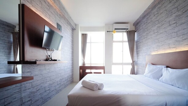 Une chambre d’hôtel avec un mur décoré d’un motif en briques, un téléviseur à écran plat fixé sur une étagère en bois, un lit avec du linge de maison blanc et une fenêtre avec des rideaux.
