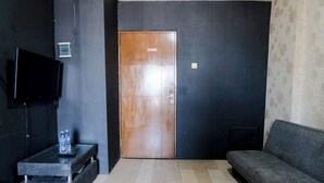 A room with a wooden door, a grey sofa, a small table with a bottle, and a TV mounted on the wall. - Tranquil Designed 2Br At Gateway Ahmad Yani Cicadas Apartment (Bandung)