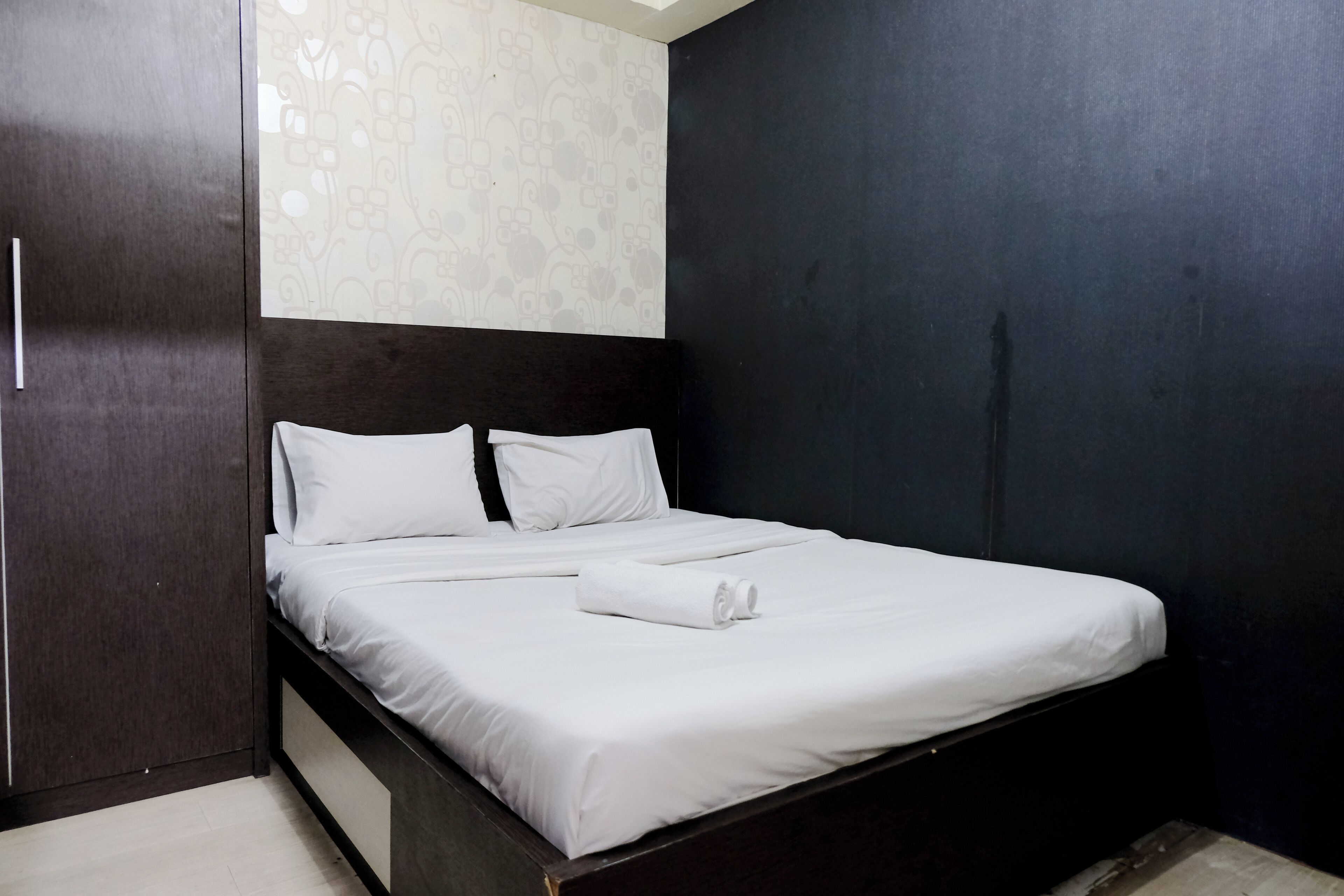 A neatly made bed with white linens and a rolled towel, a dark wooden headboard, and a wardrobe with a mirrored door.