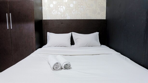 A neatly made bed with white linens and pillows, a wooden wardrobe, and a patterned wall. - Tranquil Designed 2Br At Gateway Ahmad Yani Cicadas Apartment (Bandung)