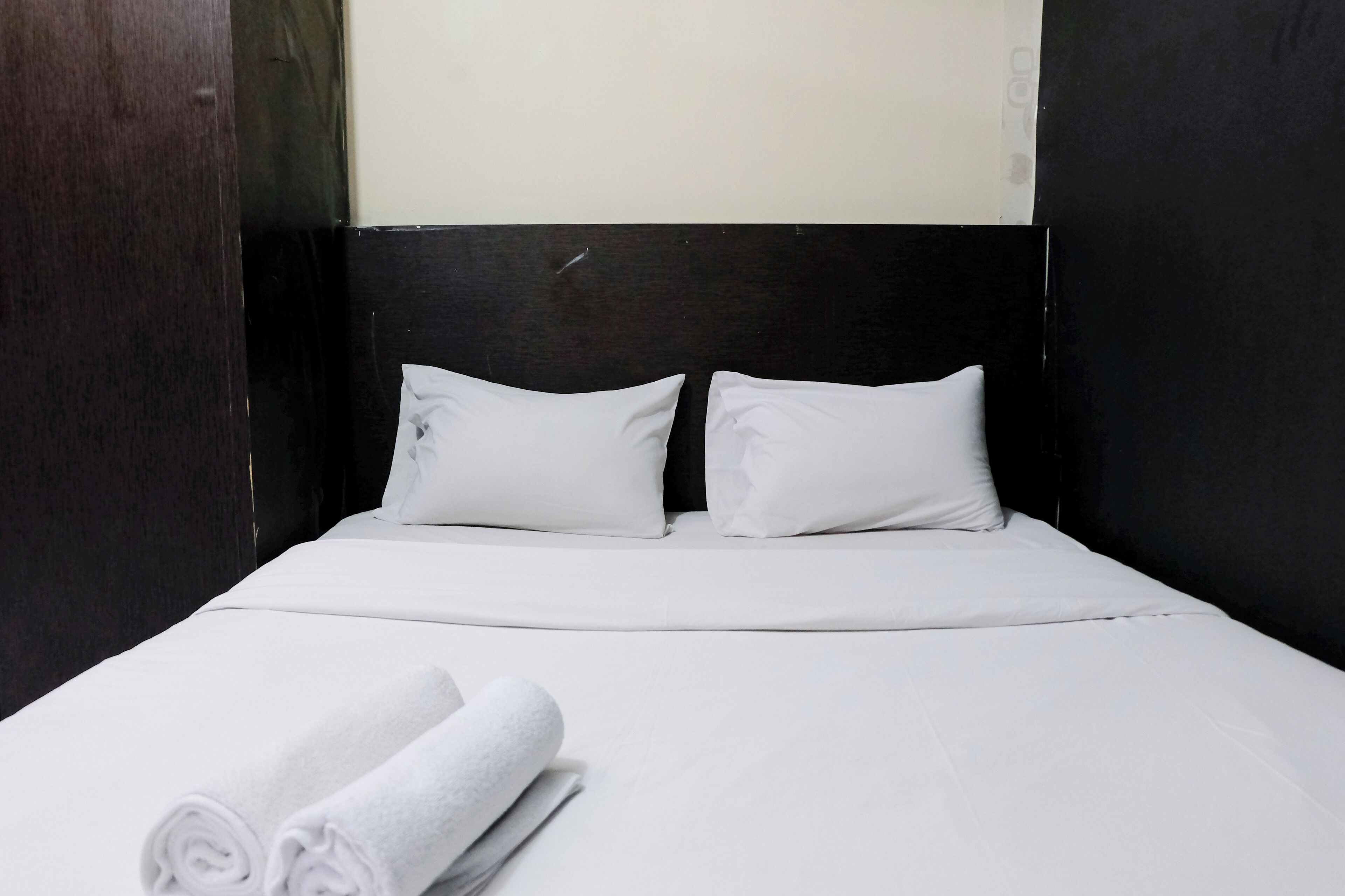 A neatly made bed with white linens and pillows, a dark headboard, and a rolled towel on the bed.