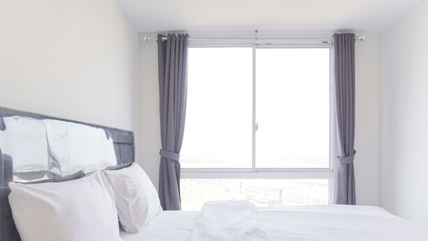 A hotel room with a bed, white bedding, and a view through a window with curtains. - Fancy And Nice 2Br Apartment At Tokyo Riverside Pik 2 (Teluknaga)