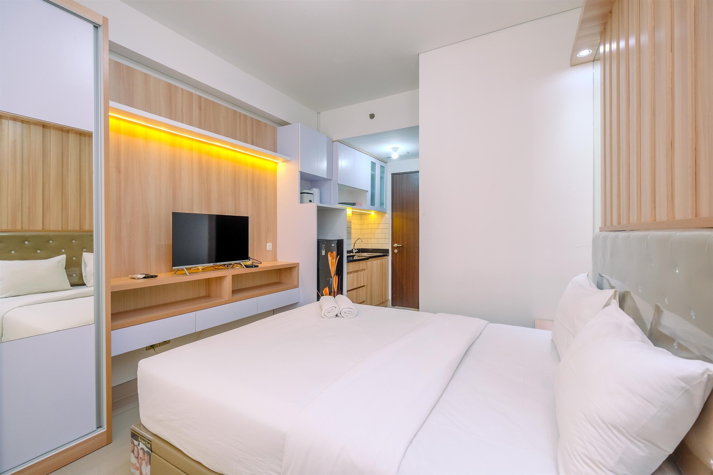 A modern hotel room with a large bed, a flat-screen TV, a built-in wardrobe, and a kitchenette in the background.
