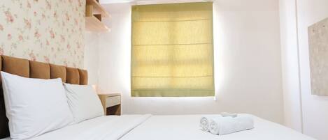 Una habitación de hotel con cama, mesita de noche, ventana con persiana amarilla y papel tapiz con motivos florales.