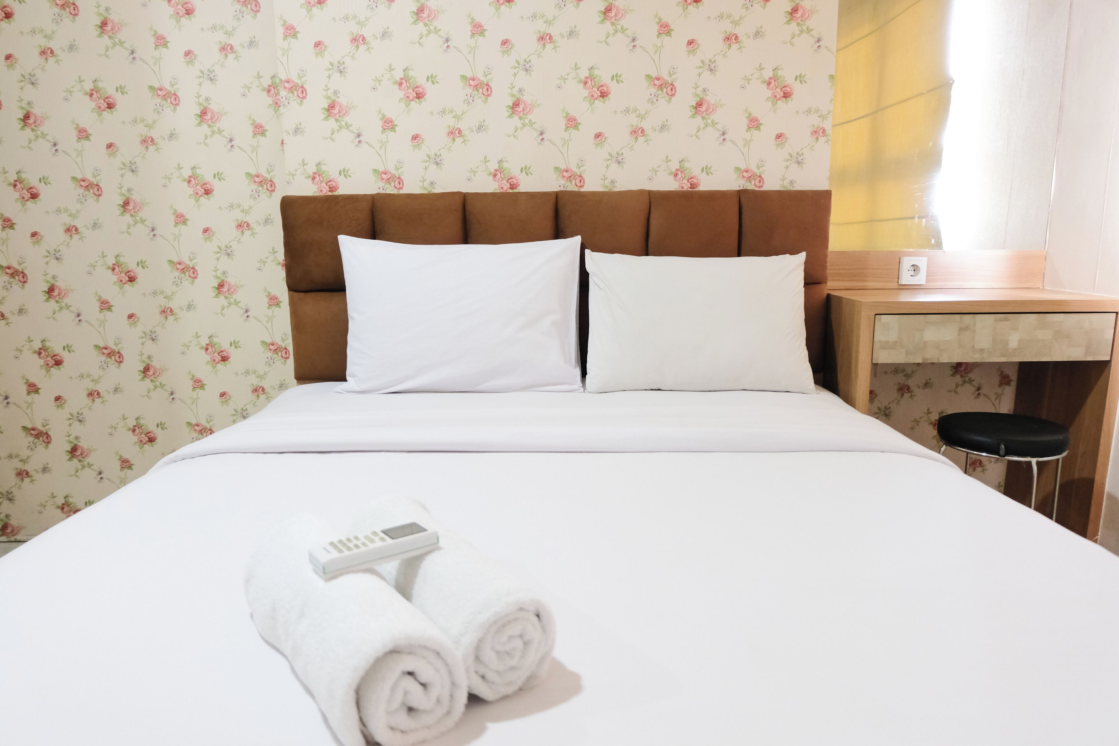 Habitación de hotel con cama, ropa blanca, cabecera, mesita de noche y un taburete pequeño.