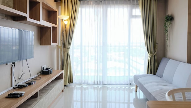 A modern living room with a flat-screen TV, wooden shelves, a sofa, and a large window with curtains. - Cozy 2Br Apartment At Gateway Pasteur (Cimahi)