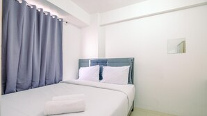 Cama bem arrumada com roupa de cama branca e toalha branca dobrada, cabeceira azul e um pequeno espelho na parede.