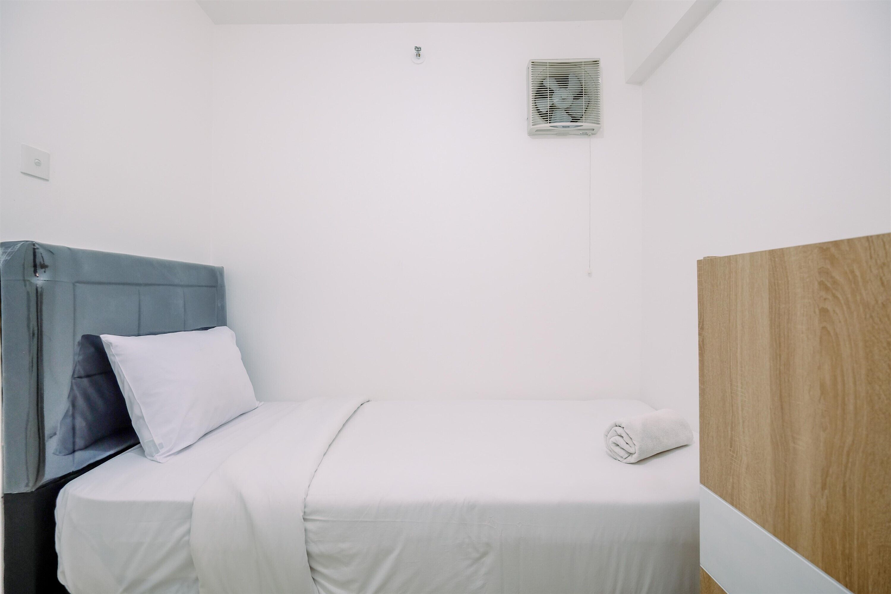 Un dormitorio pequeño y ordenado con una cama individual, cabecera azul, almohada blanca y manta blanca, además de un panel de madera en la pared.