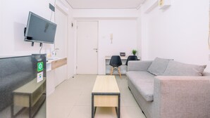 Uma sala de estar moderna com um sofá cinzento, uma mesa de madeira pequena, uma TV fixada na parede e uma escrivaninha com um computador.