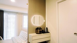 A hotel room with a bed, a bedside table, a mirror, and a small cabinet with a kettle and coffee maker. - Homey And Cozy Studio Room Daan Mogot City Apartment (Jakarta)