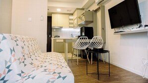 A modern living room with a patterned sofa, a dining area with two chairs, a kitchen in the background, and a mounted TV. - Homey And Cozy Studio Room Daan Mogot City Apartment (Jakarta)