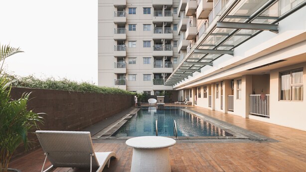 A pool area with lounge chairs and a small table, surrounded by a high-rise building with balconies. - Comfort And Homey 2Br Belmont Residence Puri Apartment (Jakarta)