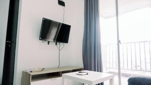 A room with a TV mounted on the wall, a small table, a wooden bench, and a view of the outdoors through a window with curtains. - Comfort And Homey 2Br Belmont Residence Puri Apartment (Jakarta)