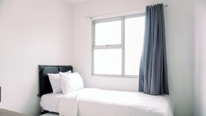 A small, neatly made bed with white bedding in a simple room with white walls and a window with a grey curtain. - Comfort And Homey 2Br Belmont Residence Puri Apartment (Jakarta)