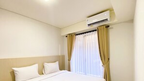 A hotel room with a bed, white linens, a window with curtains, and an air conditioning unit. - Best Homey 2Br At Meikarta Apartment (Cikarang)