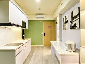 A modern kitchen with white cabinets, a microwave, and a sink. There's a wooden door with a fire safety sign, and a coffee machine on the counter. - Best Homey 2Br At Meikarta Apartment (Cikarang)