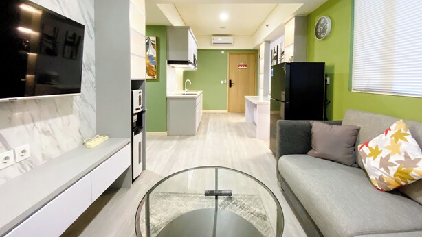 A modern living room with a glass coffee table, a grey sofa with patterned cushions, a flat-screen TV mounted on the wall, and a kitchen area with a microwave, sink, and refrigerator. - Best Homey 2Br At Meikarta Apartment (Cikarang)