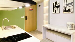 A modern kitchen with a built-in stove, a sink, and a countertop. There is a wooden door with a fire alarm sign, and abstract art on the wall. - Best Homey 2Br At Meikarta Apartment (Cikarang)