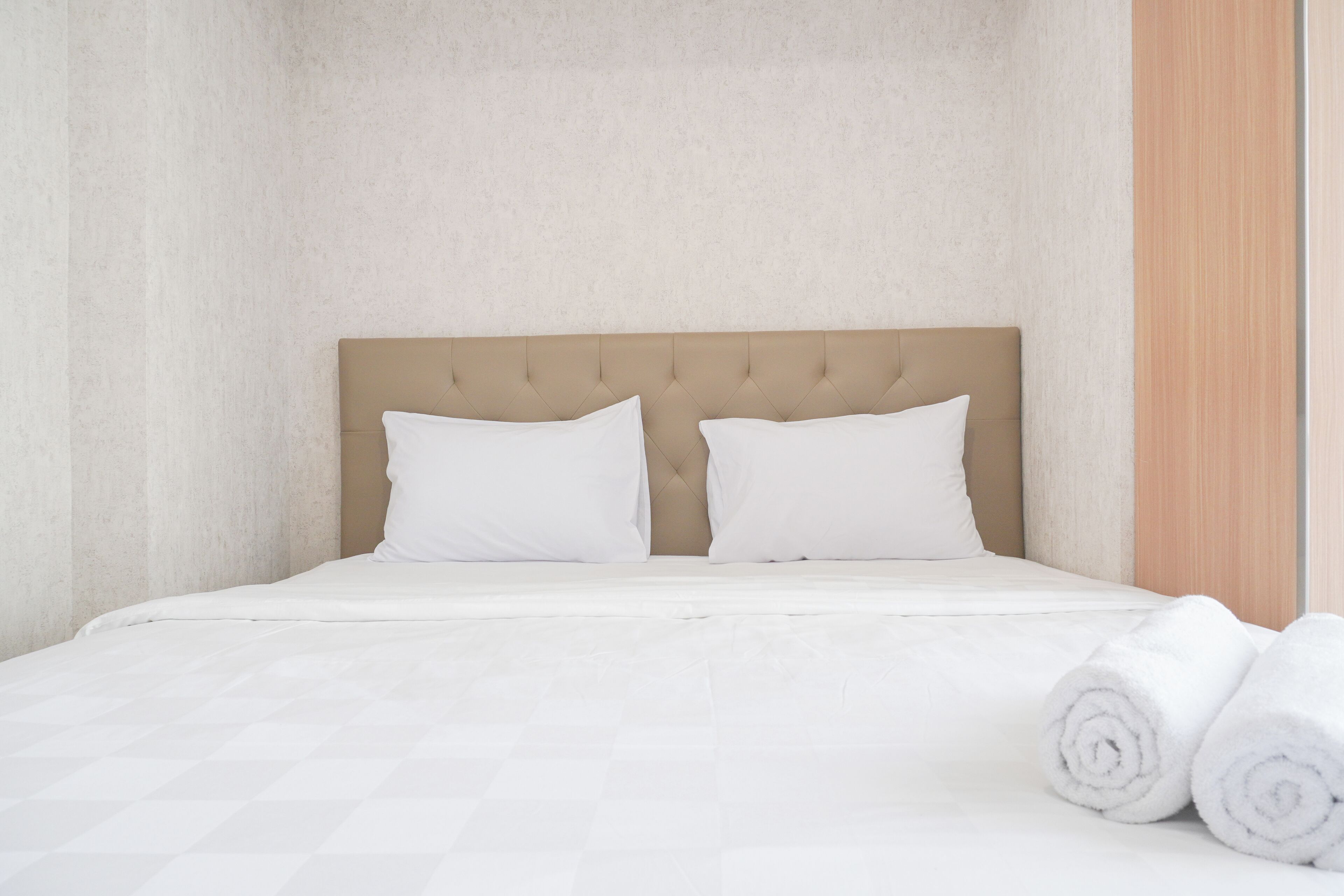 Een net opgemaakt bed met witte lakens en kussens, een beige hoofdbord en opgerolde witte handdoeken aan de zijkant.