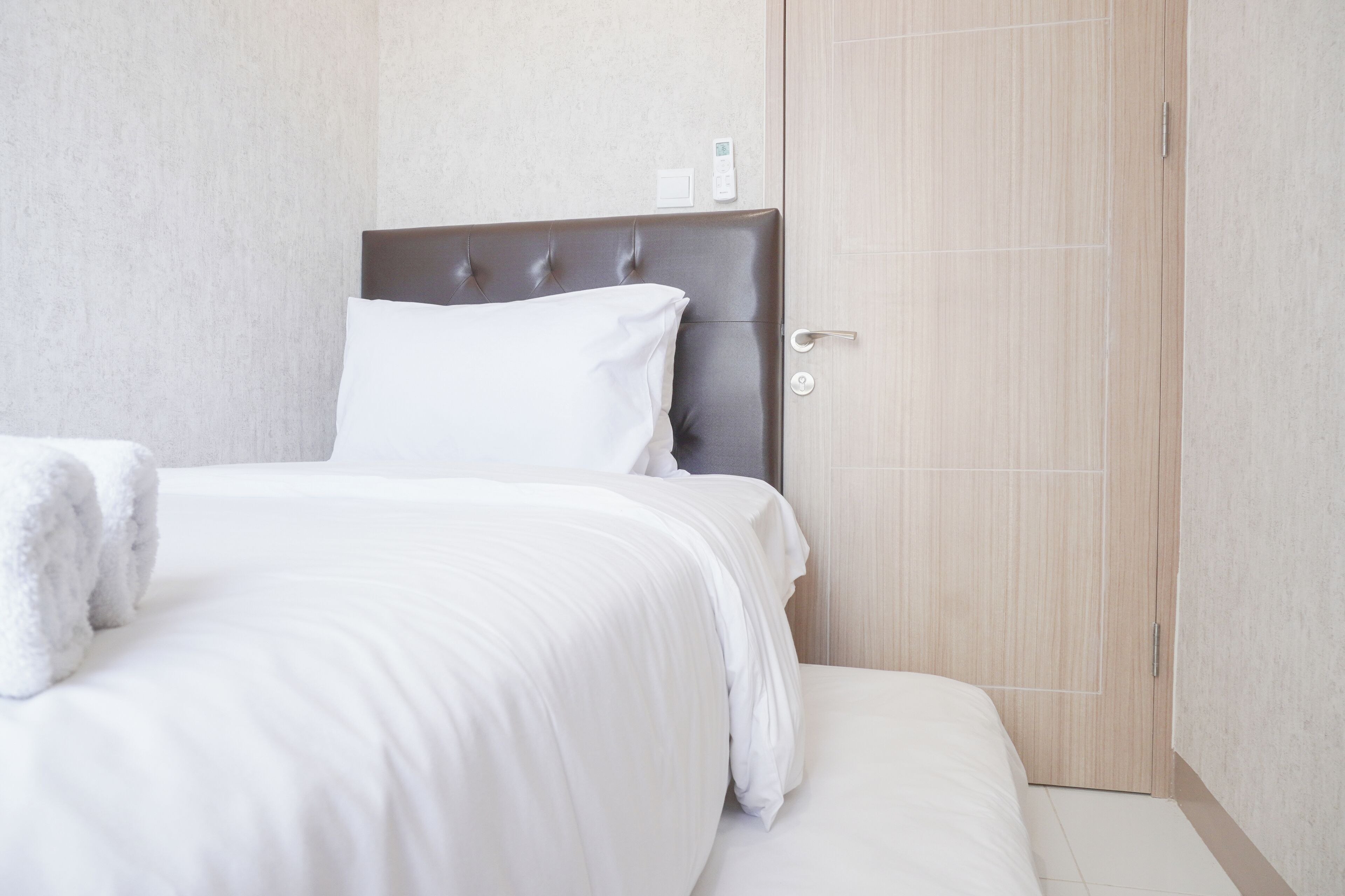 A neatly made bed with white linens and a plush headboard, a bedside table with a white lamp, and a wooden door with a handle.