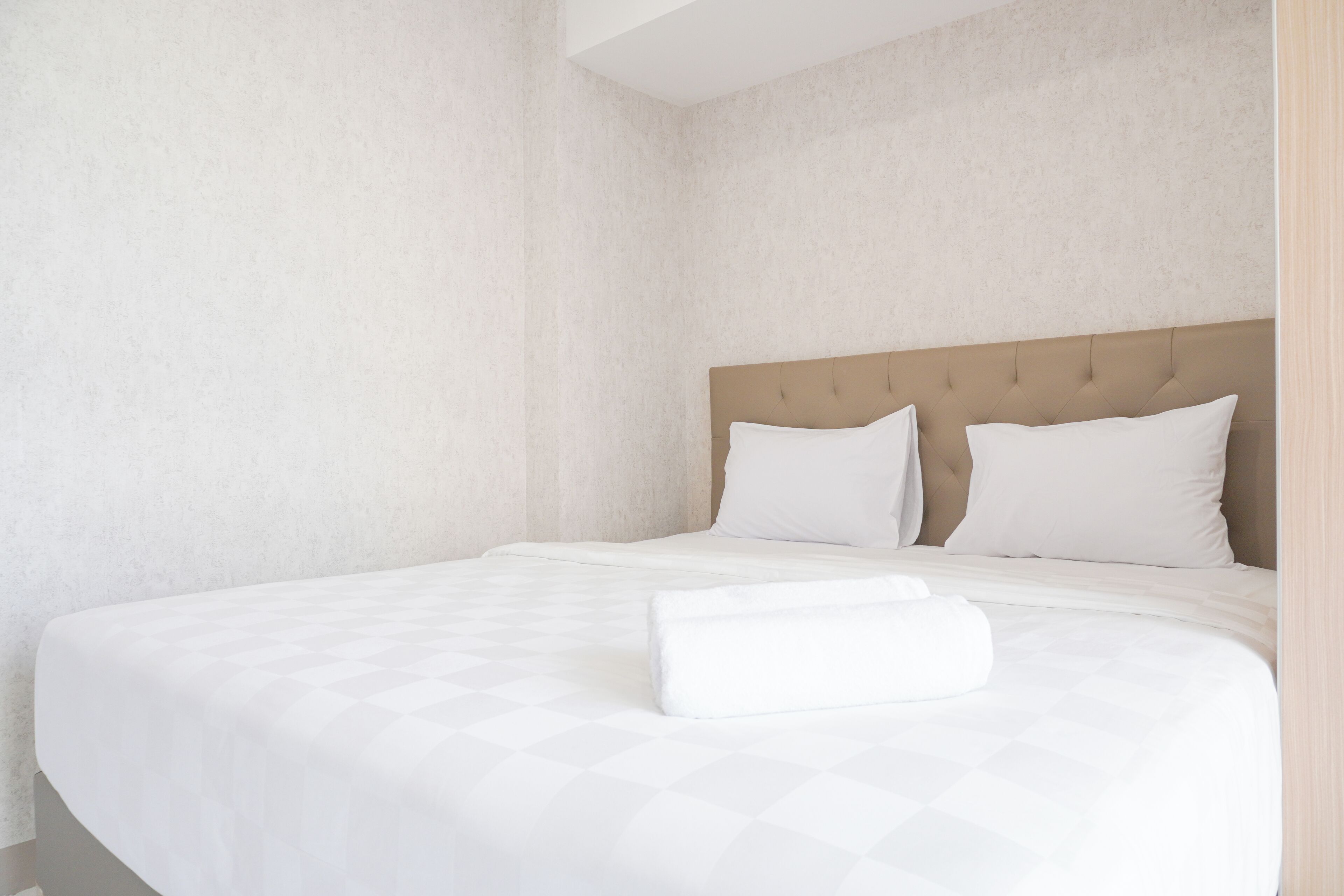 Een net opgemaakt bed met een gestoffeerd hoofdbord, witte lakens en een opgerolde handdoek op het bed.