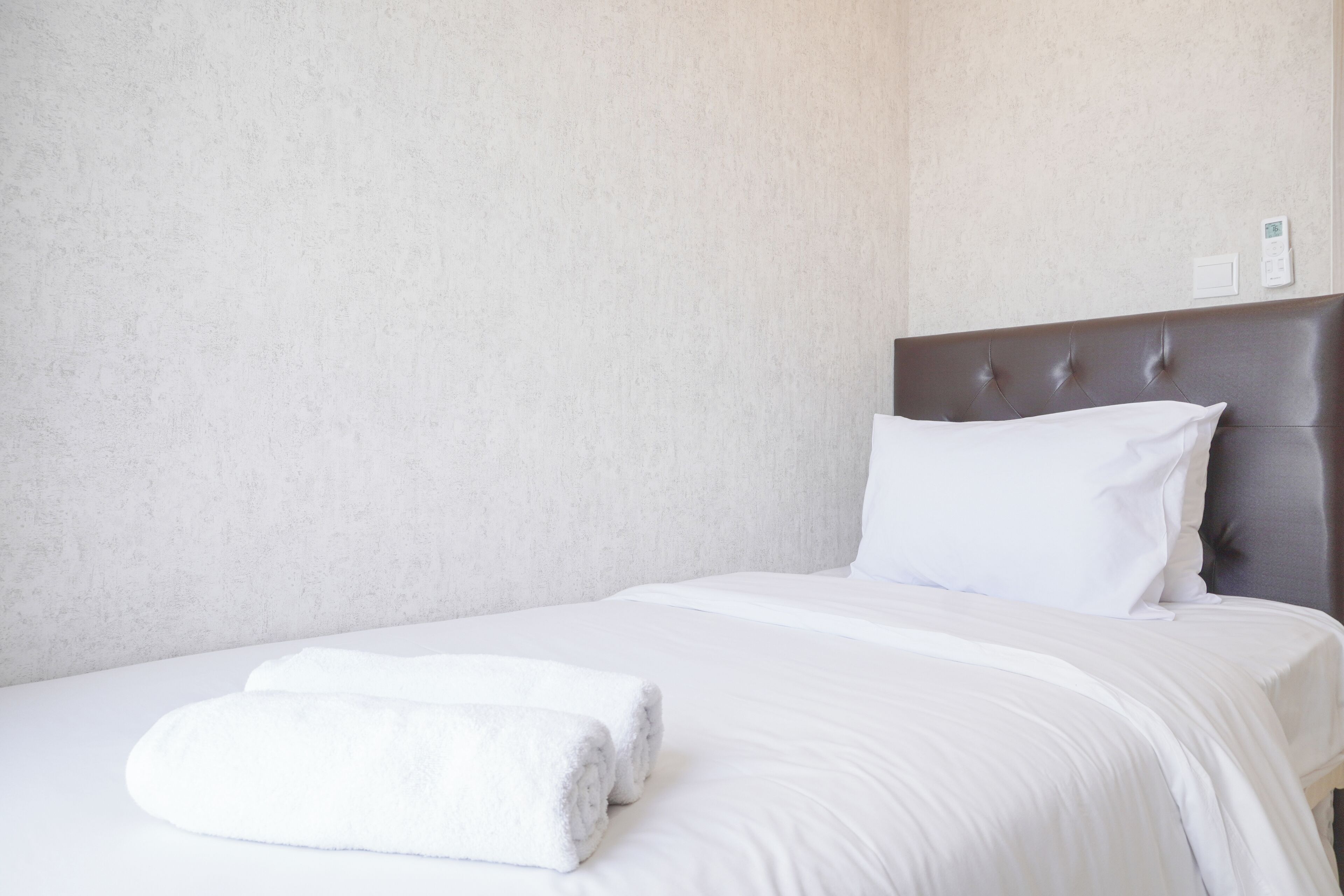 A neatly made bed with white linens and a folded white towel, a brown upholstered headboard, and a plain wall with a thermostat.