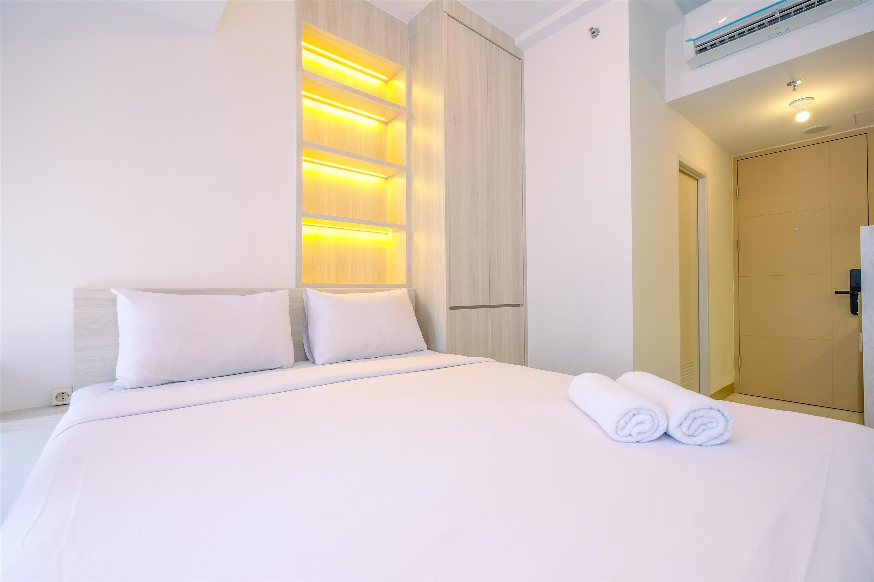 A hotel room with a neatly made bed, a built-in shelf with lighting, and a door to the right.